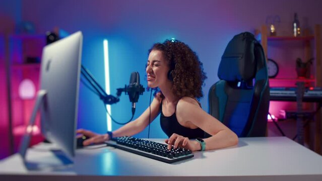 Young woman wearing headphones playing computer game neon fashion room, winner. Female gamer girl blogging, commenting hard match, looking at computer monitor, using computer mouse and keyboard