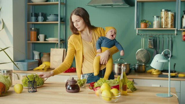 Tired Mom SoothesThe Child on the Hands Trying Makes Salad.Housewife Multitasking at Home With Her Newborn Baby.Woman Housewife Holding Infant and Cooking Dinner in Kitchen
