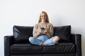 Obraz premium Happy young woman using mobile phone while sitting a couch at home with laptop computer