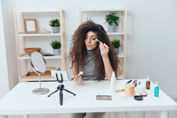 Makeup school concept. Professional visage artist tanned handsome curly Latin beauty blogger in casual top in home interior. Copy space Banner. Influencer record video review blog using smartphone