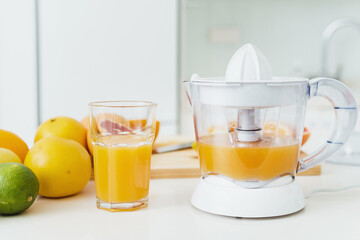 Glass of orange juice, citrus juicer and various citrus fruits on kitchen table