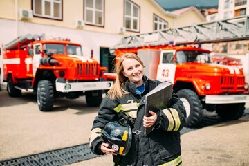Female firefighter in in a protective suit with documents standing ain the background of a fire engines. Fire station