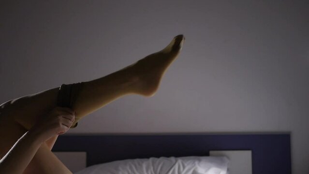 Close-up of female leg in erotic beige stockings. Young caucasian woman putting stocking on her leg lying on the bed.