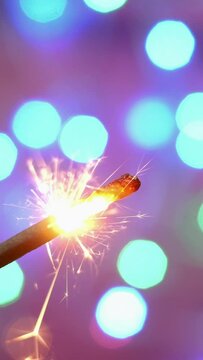 Bright burning sparkler against backdrop of defocused multicolored blinking garland. Blurred backdrop. Merry Christmas or birthday holiday concept.