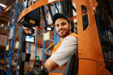 Happy male worker driving forklift at distribution warehouse and looking at camera. © Drazen