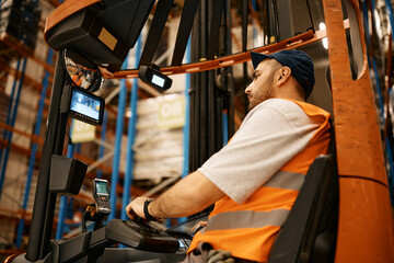 Young man driving forklift while working at storage compartment of distribution warehouse.