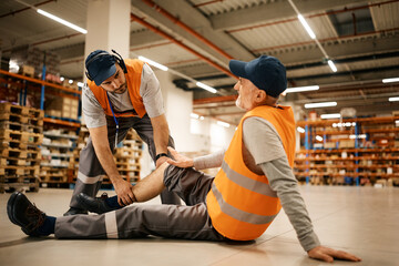 Young worker assists his colleague with leg injury while working at distribution warehouse. - obrazy, fototapety, plakaty