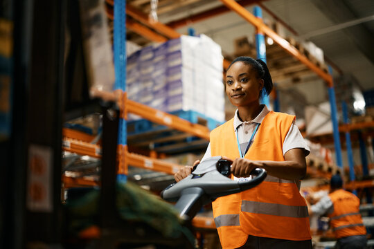 Black female worker pushing cargo on pallet jack while working at distribution warehouse.