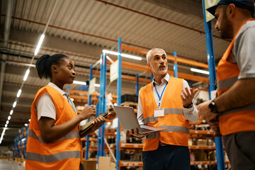 Mature manager using laptop while talking with workers at distribution warehouse.