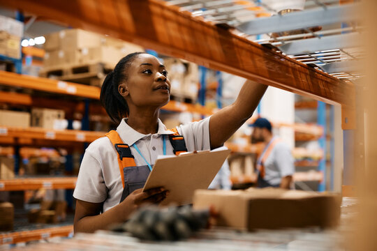 Black female worker checking inventory on shelves of distribution warehouse.