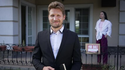 Portrait of satisfied seller looking at camera smiling with blurred buyer standing on house porch at background. Front view Caucasian man posing in slow motion selling real estate property