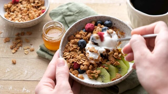 Bowl of crunchy oat honey granola with yogurt and berries. POV female hands eating granola bowl, healthy breakfast food