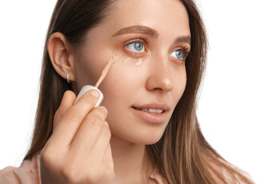 Close up photo of a concentrated woman applying concealer while looking up. Studio shot over white background.