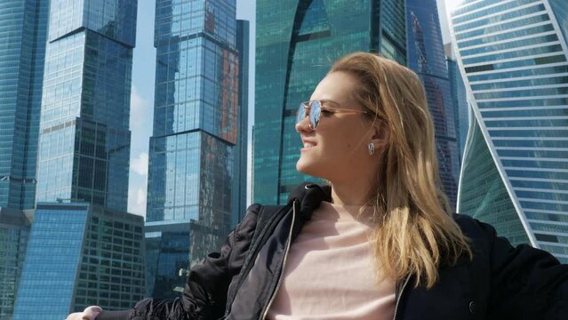 Portrait of beautiful blonde girl in sunglasses. Pretty lady smiling against of skyscrapers with mirrored walls. Young woman admiring views of metropolis near glass highrises. Daytime