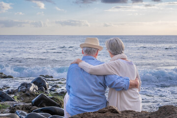 Rear view of relaxed caucasian senior couple sitting on the pebble beach at sunset light admiring horizon over water. Two gray haired elderly people hugging with love