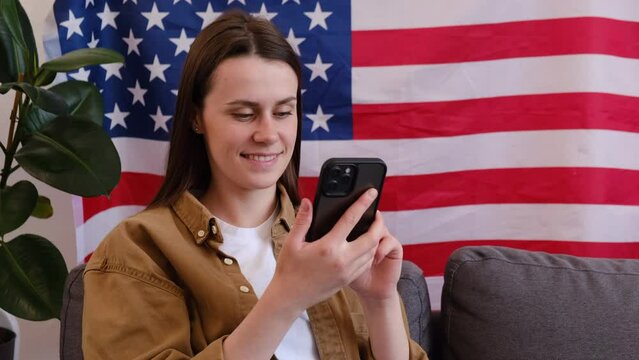Beautiful smiling young brunette woman sitting on cozy sofa at home with USA flag, cute happy female 20s old years with smartphone looking Independence Day celebration. Patriotic US holiday concept