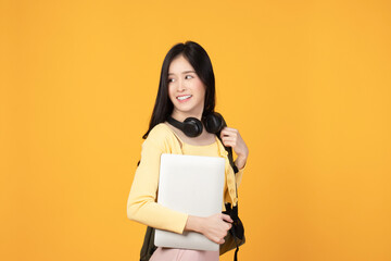 Portrait of happy casual Asian girl student with backpack, headphones and laptop isolated on yellow background. Back to school and learning concept.