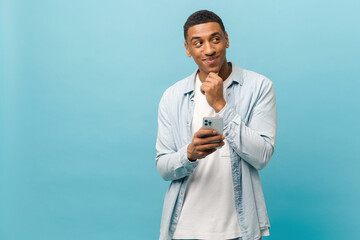 Thoughtful young multiracial short-cutted man in casual shirt standing with smartphone and holding...