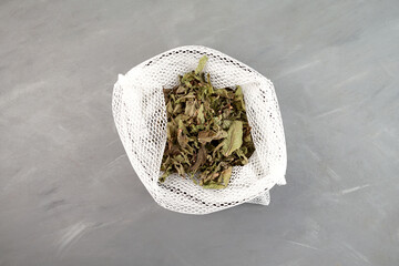 Dried melissa in in mesh eco-bag for storing, top view. Leaves for making herbal tea. Grey background, selective focus - obrazy, fototapety, plakaty