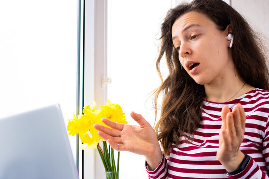 Beautiful young woman talking with client or partner on video call conference using laptop near window indoor. Discussing project details online, working distant remotely from home using 5g internet