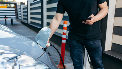 Young man traveling by electric car at summer, having stop at charging station standing plugging cable and browsing internet on smartphone joyful while charing