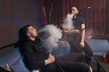 young men smoke a hookah, sitting on a sofa and exhaling thick white smoke. in the foreground a bowl of hookah on grapefruit