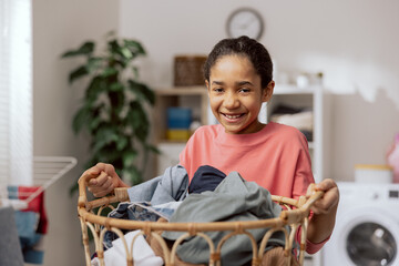 Smiling pretty girl stands in the middle of the bathroom, laundry room, holding a large wicker basket filled with colorful clothes in hands, daughter helps mother with household chores.