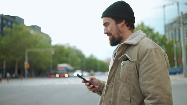 Serious bearded man wearing hat walking and holding his mobile in the city 