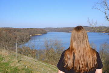 A girl is resting on a clear day on the shore of a blue lake with wooded hilly shores in early spring. View from the back.