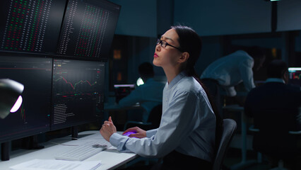 Asian woman trader look at multi-screen workstation with graphs and data working late in dark office