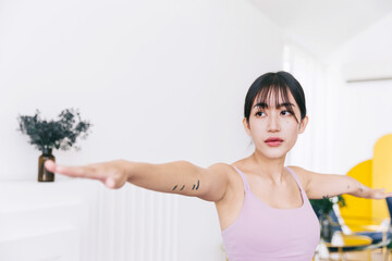 Young smiling attractive sporty Asian woman practicing yoga, doing Virabhadrasana 2 exercise, meditating in Warrior Two yoga pose, indoor working out at home, wearing sportswear. Half-length photo.