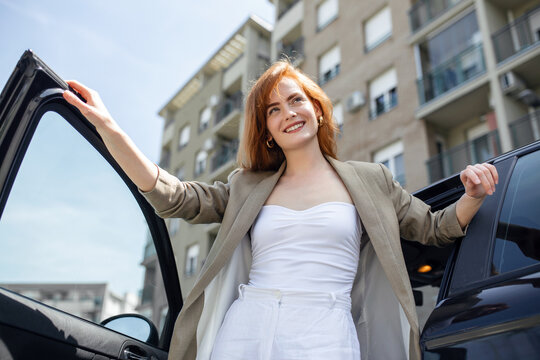 Stunning young woman in entering her car holding the door with one hand and leaning on the hood with the other