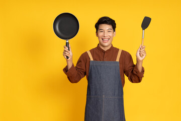 Young Asian man chef wearing kitchen apron cooking and holding pan and spatula isolated on yellow background