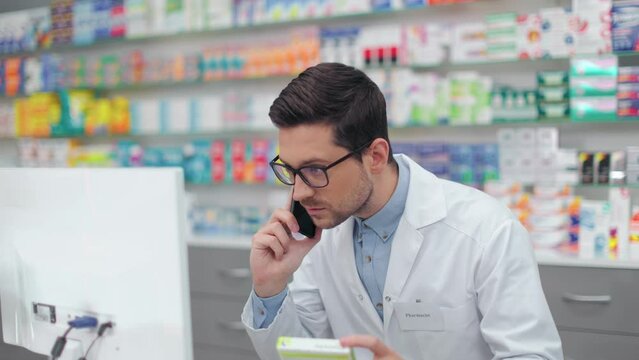 Competent male pharmacist in eyeglasses and lab coat provision consultation about pills to client during mobile conversation. Advice, technology and medical care concept.