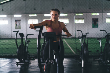 Naklejka na ściany i meble Portrait of determined woman training on exercising bike reaching dedication results during intensive cardio training, young female slimming in modern gym - keeping healthy lifestyle and vitality