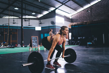 Obraz na płótnie Canvas Young female in active sportswear pick up barbell equipment during weightlifting training practice in gym studio, strong fit girl have deadlift workout for keeping body muscles in tonus - stamina