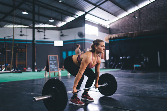 Young female in active sportswear pick up barbell equipment during weightlifting training practice in gym studio, strong fit girl have deadlift workout for keeping body muscles in tonus - stamina