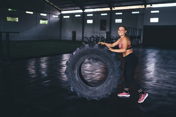 Obraz na płótnie Canvas Portrait of cheerful female in active wear using wheel tire for slimming and working on arms muscles in gym, happy professional sportive woman having hard cardio workout for training strength