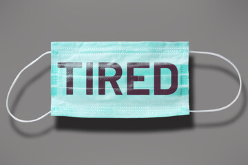 Blue disposable mask with word: TIRED. Covid routine prevention. Abstract concept of seasonal epidemic safety against virus spreading.