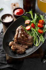 Grilled beef steak, well done with fresh vegetable salad and a glass of light beer on a black plate on the dark gray background top view