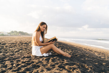 Fototapeta na wymiar Beautiful female tourist in casual clothes reading literature best seller during leisure pastime an sandy coastline beach, Caucaisan woman interested in novel poetry book - resting at seashore