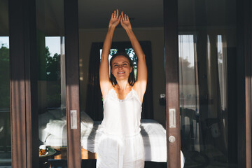 Half length portrait of cheerful female in silk nightgown smiling at camera while stretching after...