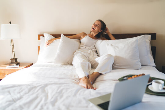 Joyful Caucasian blogger with laptop technology laughing during leisure time in hotel bedroom, cheerful female freelancer with digital netbook technology enjoying room service - rejoices and delight