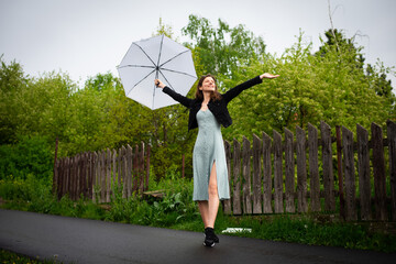 A happy young woman in a light delicate dress walks with a light umbrella in rainy weather.