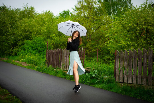 A young beautiful woman in a light delicate dress walks with a light umbrella in rainy weather.