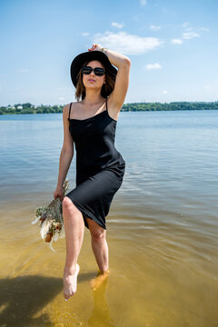 Pretty woman walking along barefoot in evening black dress in city lake
