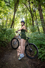 Fototapeta na wymiar Young slim woman in green dress and hardhat for safety stand near bike on park