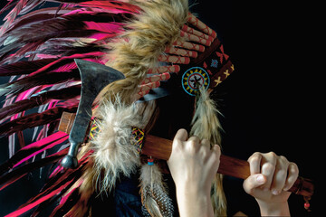 A young girl in an Indian roach holds a tamahawk in her hands near her head on a black background.