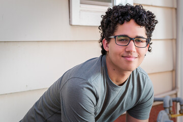 A young man with curly hair poses in front of a beige siding wall.