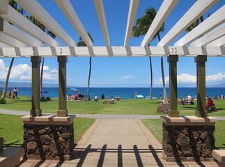 View of a sunny beach from a white wooden shelter. Symmetrical shadows opposite to the roof. Deep blue sea, clear blue sky, palm trees, green lawn, stone pillars, people sunbathing and relaxing.